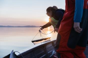 A person releases a fishing net from a boat at sunset with another person nearby.